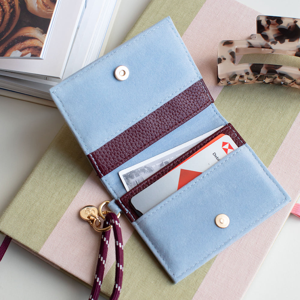 A light blue suede wallet with burgundy faux-leather trim, shown open with cards in the slots and a matching burgundy-and-white wristlet strap attached. The wallet is photographed on a pastel striped notebook with a hair clip and framed photo in the background.