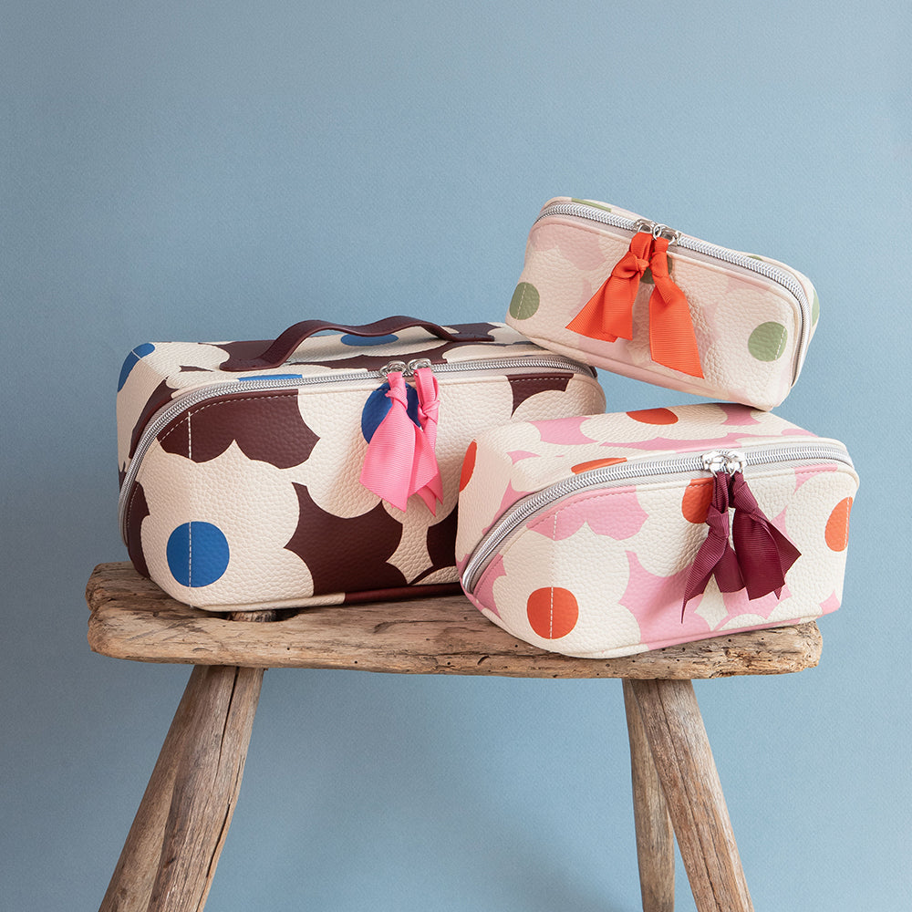 Three patterned zippered cosmetic pouches in different sizes displayed on a rustic wooden stool against a soft blue background. The pouches feature a cream base with large brown, pink, orange, and blue floral-like shapes, along with bright orange, pink, blue, and burgundy tassels.