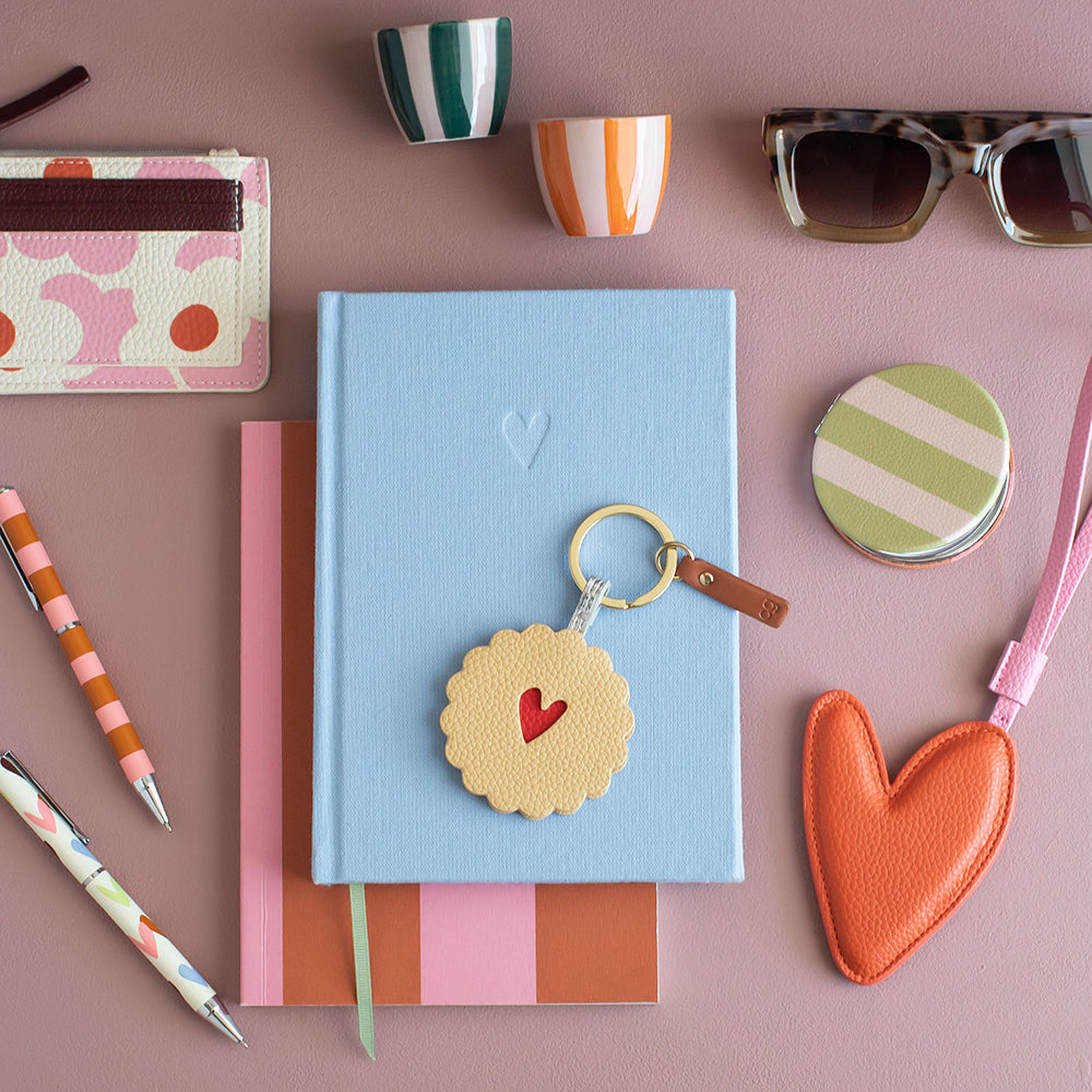 A styled flat-lay featuring a light blue textured notebook with a round beige leather keychain charm shaped like a sliced cookie with a red heart, surrounded by coordinating stationery and accessories on a dusty pink background.
