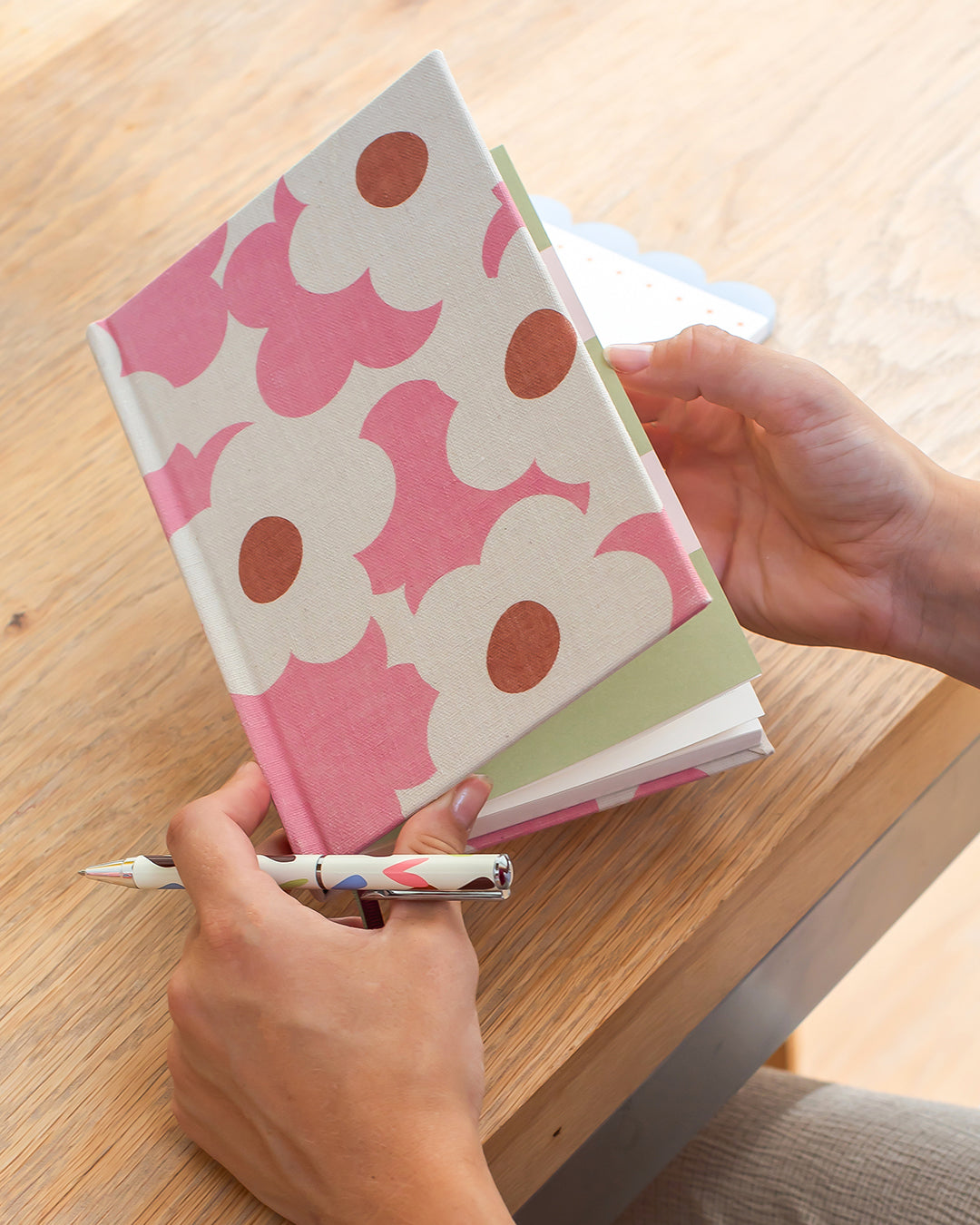 A person holds a hardcover notebook with a pink and white floral pattern and a matching patterned pen on a light wood table.