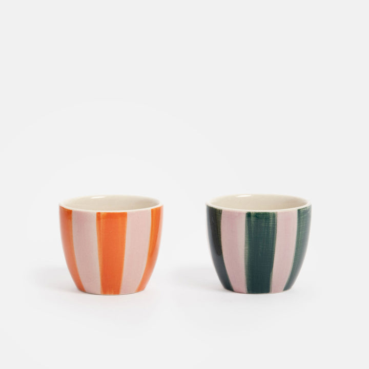 Two small ceramic egg cups with vertical stripes, one in orange and light pink, and the other in dark green and light pink, set against a plain white background.