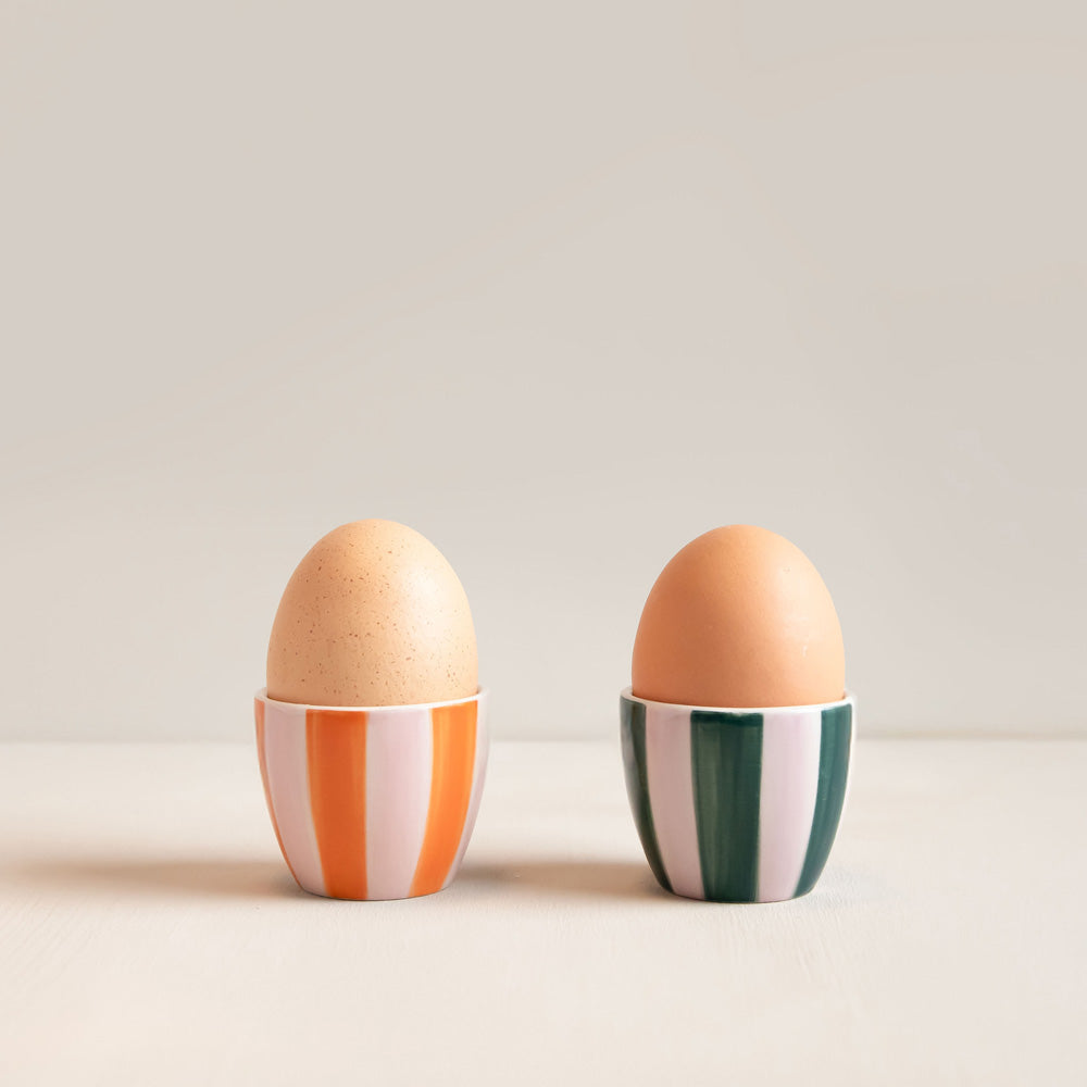 Two ceramic egg cups holding brown eggs, set against a minimalist light beige background. The egg cup on the left features vertical orange and white stripes, while the egg cup on the right features vertical dark green and white stripes.