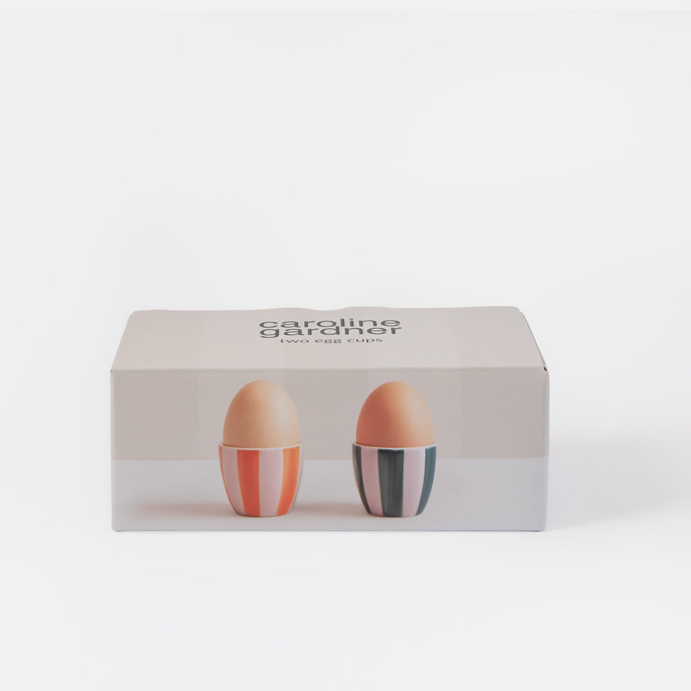 Caroline Gardet Egg Cup Set in its original packaging, featuring two ceramic eggs in orange, pink, and gray striped eggshells, displayed on a clean white background.