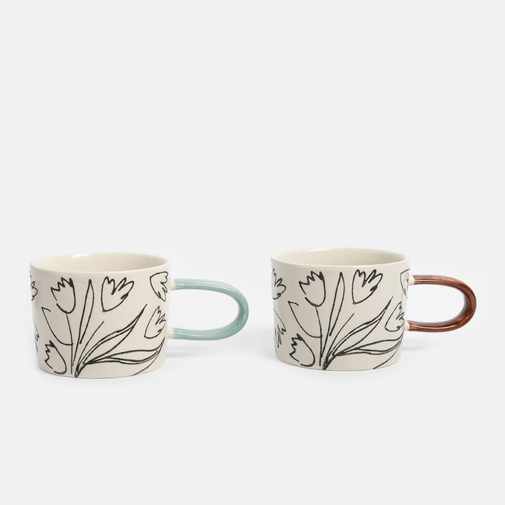 Two ceramic mugs with a black tulip line-art pattern on a warm off-white background, one with a light blue handle and the other with a copper-brown handle, set against a light gray studio background.
