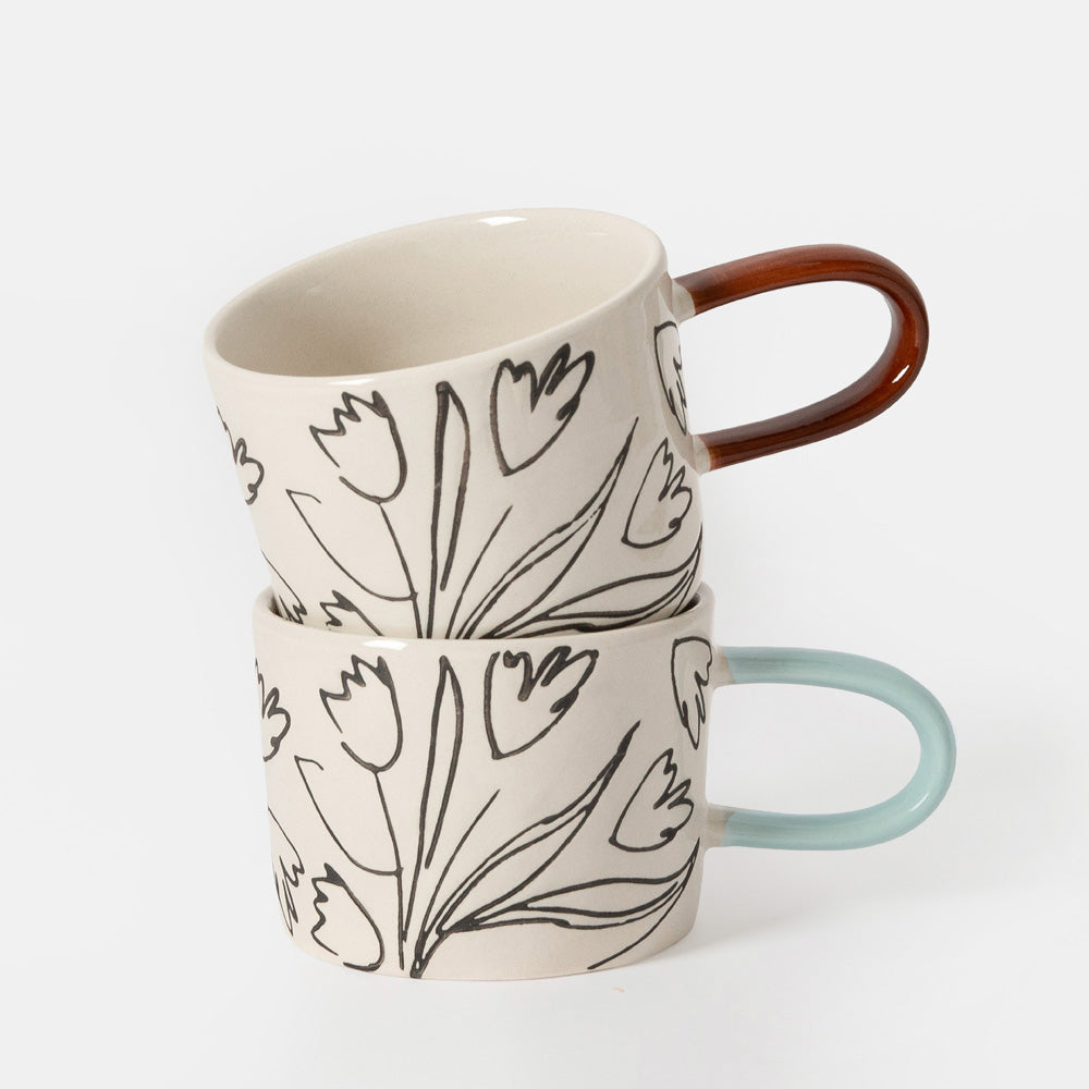 Two ceramic mugs with a black tulip line-art pattern on a warm off-white background, one with a light blue handle and the other with a copper-brown handle, set against a light gray studio background.