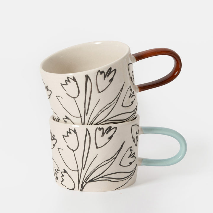 Two ceramic mugs with a black tulip line-art pattern on a warm off-white background, one with a light blue handle and the other with a copper-brown handle, set against a light gray studio background.