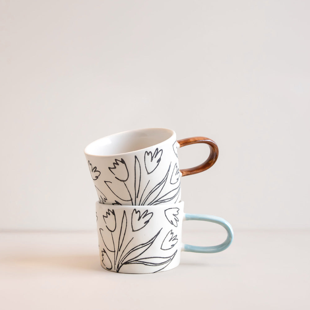 Two stacked ceramic mugs with a black tulip line-art pattern on a white background. The top mug has a brown wooden handle, and the bottom mug has a light blue handle.