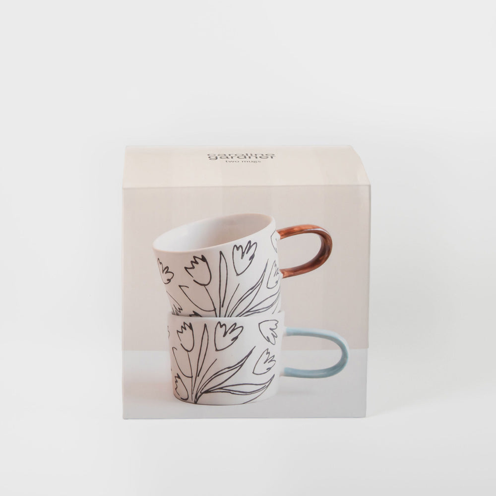 A boxed set of two stacked ceramic mugs with a white background and black floral line drawings. The top mug has a copper-brown handle, and the bottom mug has a light blue handle.