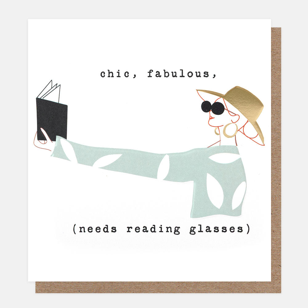 A square white greeting card with a minimalist illustration of a woman in a wide-brimmed hat and reading glasses, reading a book, with the text 'chic, fabulous, (needs reading glasses)'. A brown kraft envelope is partially visible behind the card.