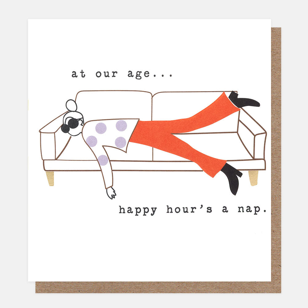 A white greeting card with a minimalist illustration of a person lying on a couch, wearing bright orange pants and a white top with purple polka dots. The card has black text reading 'at our age...happy hour's a nap'.