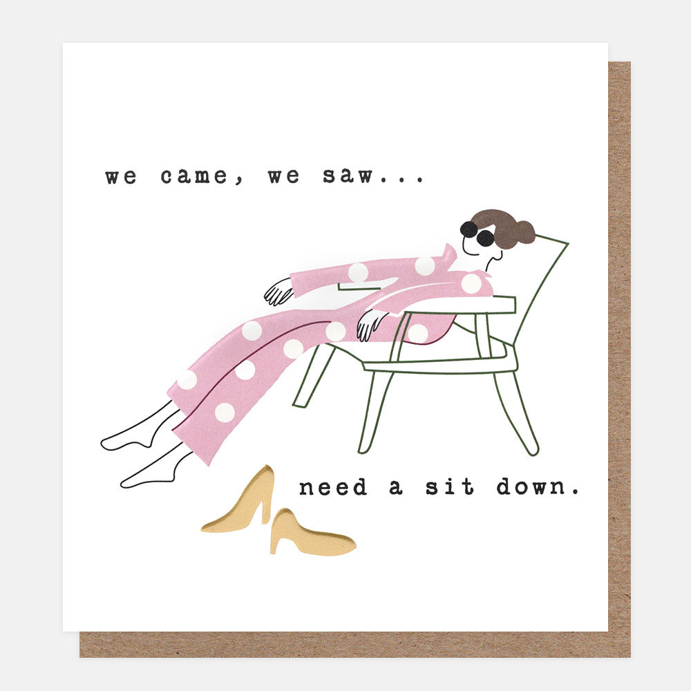 A white greeting card with a brown kraft envelope behind it. The card features a minimalist line drawing of a woman lounging in a chair wearing pink polka-dot pajamas and sunglasses, with the text 'we came, we saw...need a sit down'.
