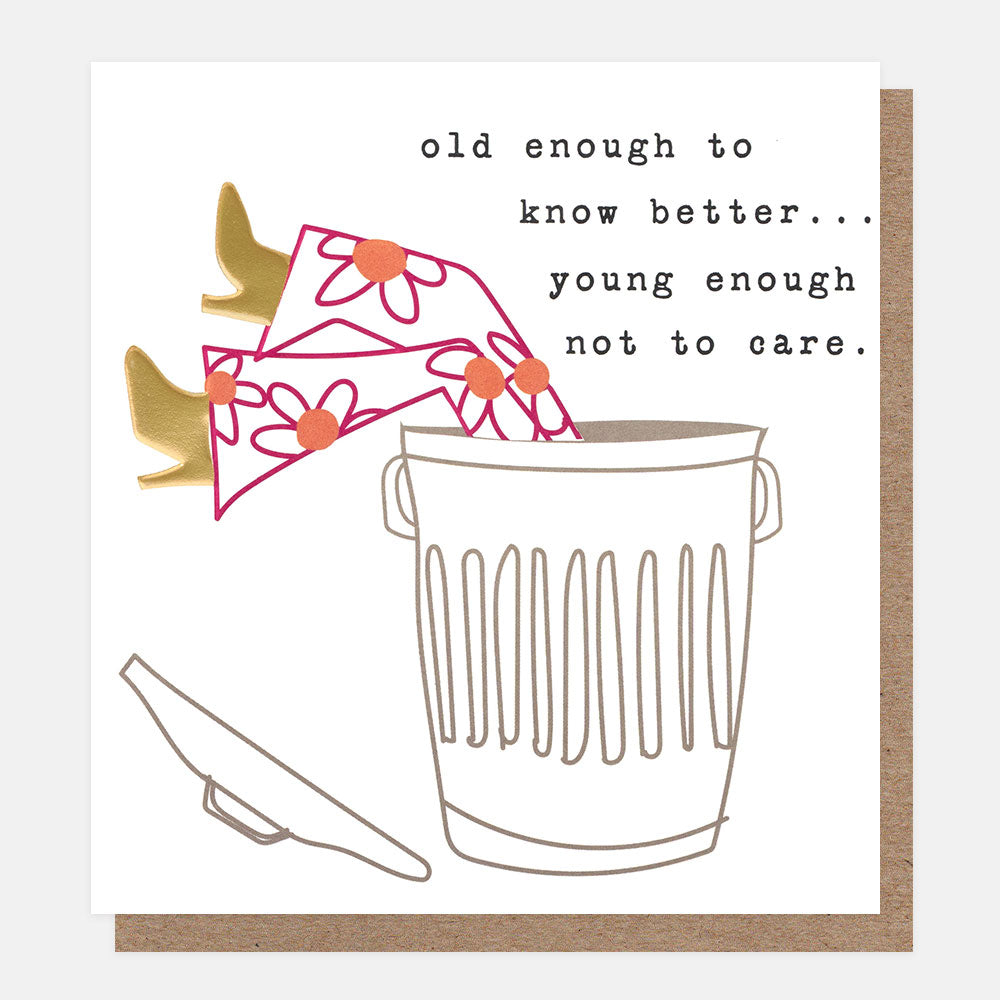 A square greeting card with a white front and a brown kraft envelope behind it. The card features a minimalist line drawing of a woman slumped in a dustbin with her legs and heels sticking out of the top, with the text, 'old enough to know better...young enough not to care'.