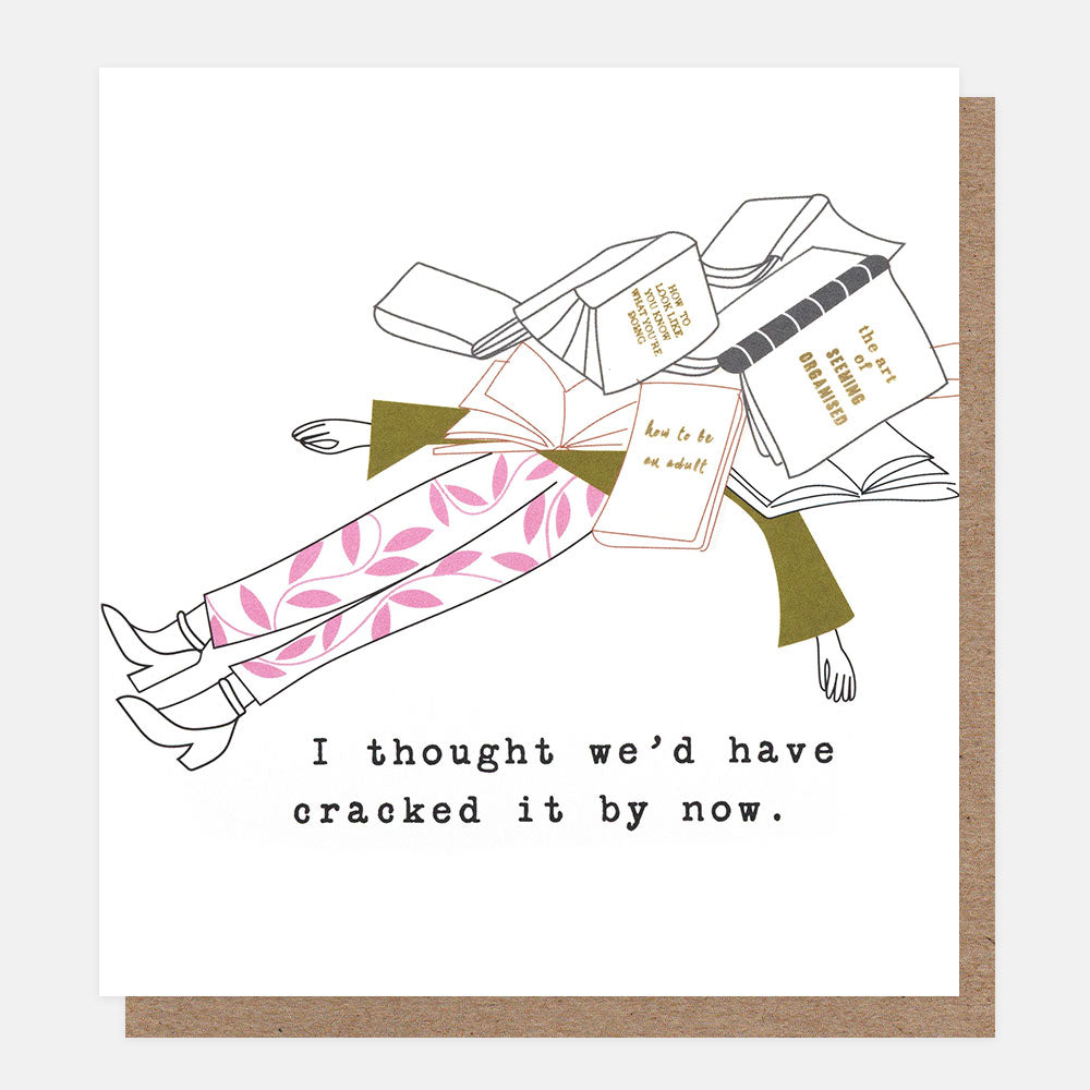 A white greeting card with a minimalist line-art illustration of a person lying on their back surrounded by books, with pink patterned pants and olive green sleeves. The card has black text at the bottom that reads, 'I thought we'd have cracked it by now'.