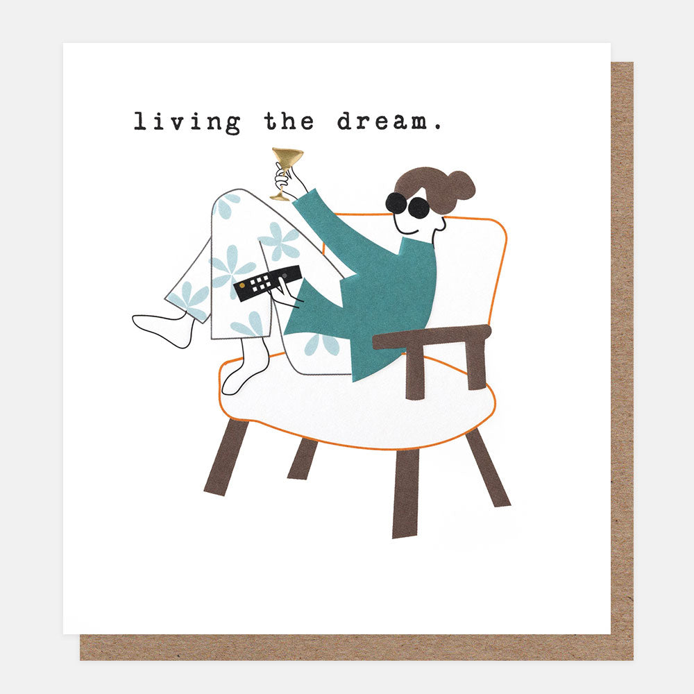 A square greeting card with a white front and a brown kraft envelope behind it. The card features a minimalist illustration of a person lounging in an armchair, holding a drink and sunglasses, with the text 'living the dream'.