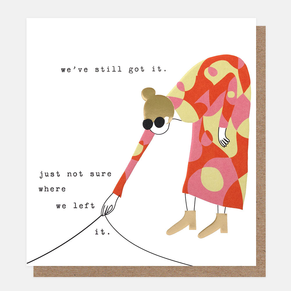 A square greeting card with a white front, featuring a stylized illustration of a person in a red, pink, and yellow patterned dress bending down and looking under a rug. Black text on the card reads, 'we've still got it. just not sure where we left it'.