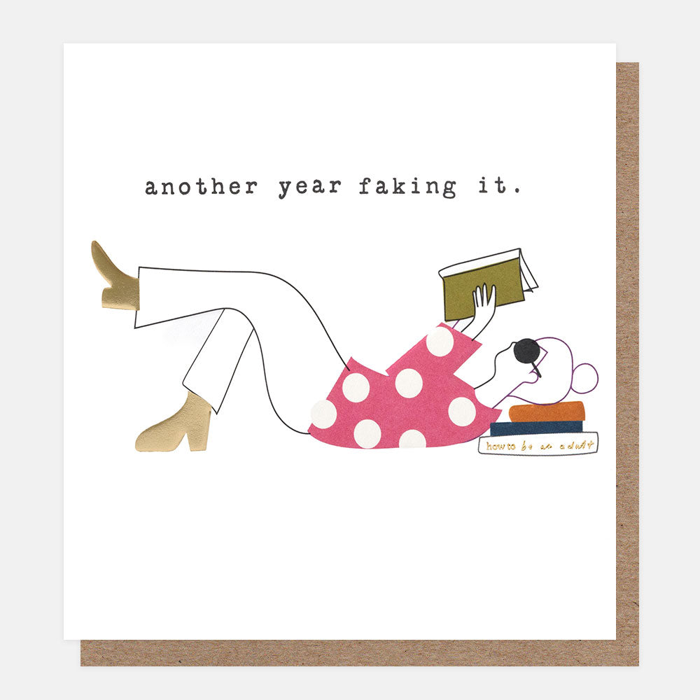 A square white greeting card with a brown kraft envelope behind it. The card features a minimalist line drawing of a person in a pink polka-dot dress reading, with the text 'another year faking it.