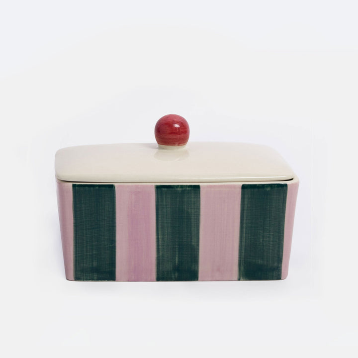 A rectangular lidded ceramic butter dish  with a cream-colored top and lid, featuring vertical stripes in dusty pink and dark green on the sides, topped with a small round red knob, set against a plain white background.