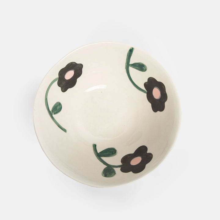 An off-white ceramic bowl with a hand-painted floral pattern of black flowers, green stems, and pink centers, shown from a top-down view on a light gray background.
