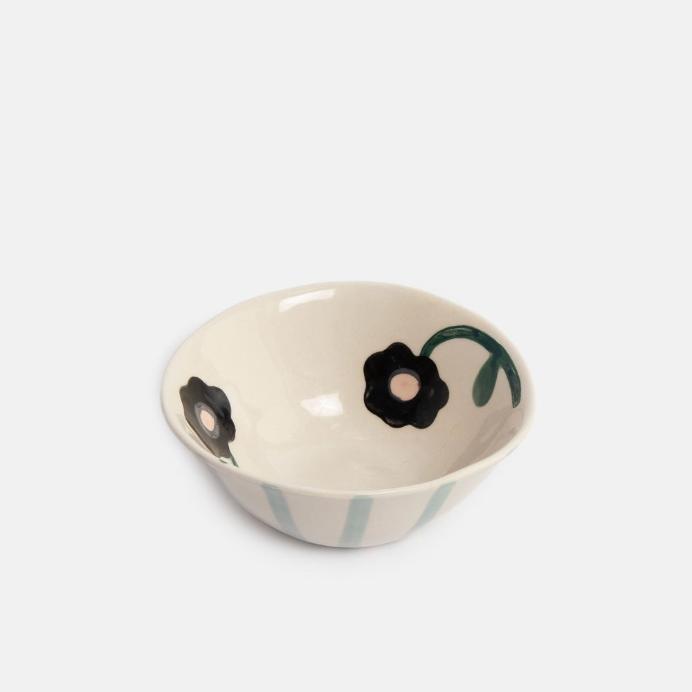 A small ceramic bowl with a cream-colored interior decorated with black flowers and green stems, and light blue vertical stripes on the exterior, set against a plain light gray background.