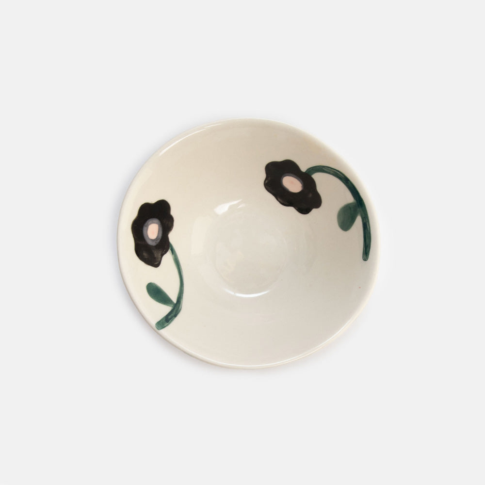 An off-white ceramic bowl featuring a hand-painted floral pattern with black flowers and green stems, shown from a top-down perspective on a light gray background.