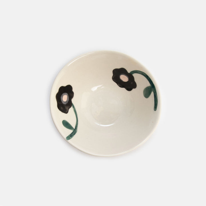 An off-white ceramic bowl featuring a hand-painted floral pattern with black flowers and green stems, shown from a top-down perspective on a light gray background.