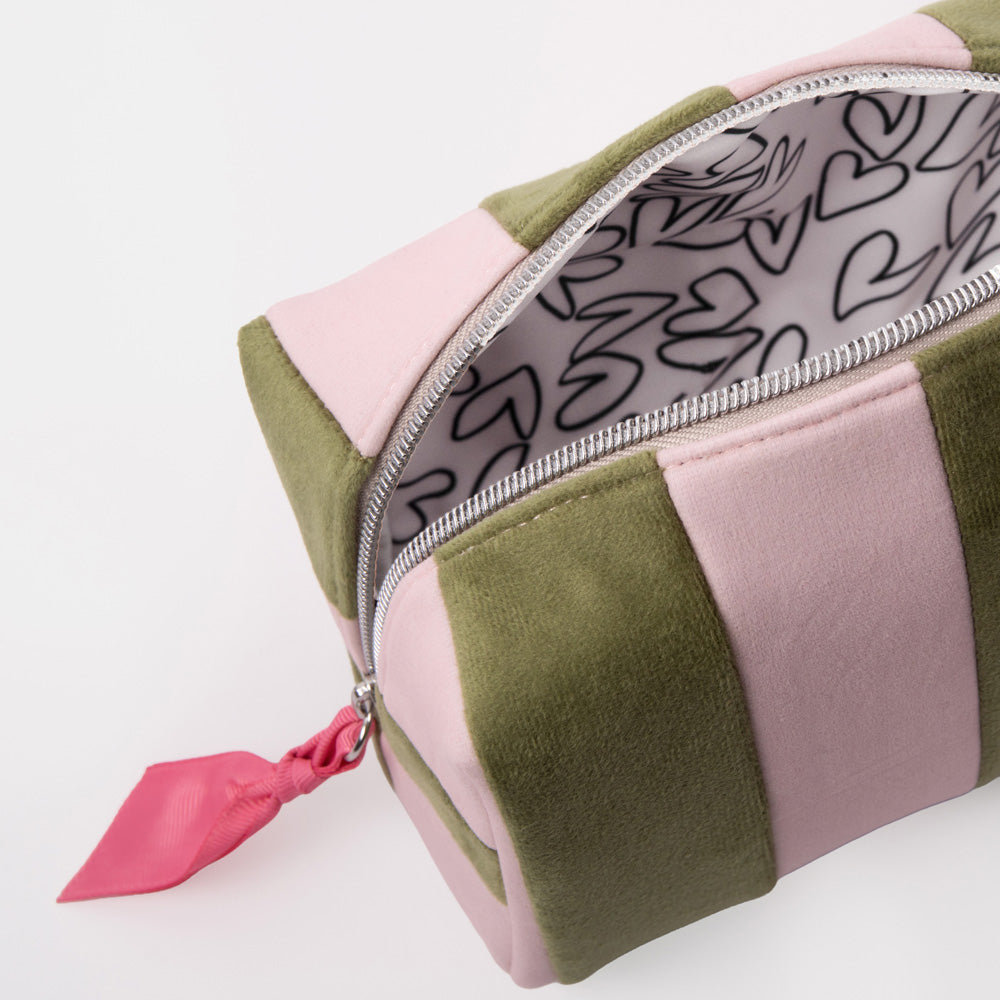 A close-up of a rectangular zippered pouch with alternating light pink and olive green velvet-like panels, a silver zipper, and a bright pink pull tab. The inside lining has a white background with black heart outlines.