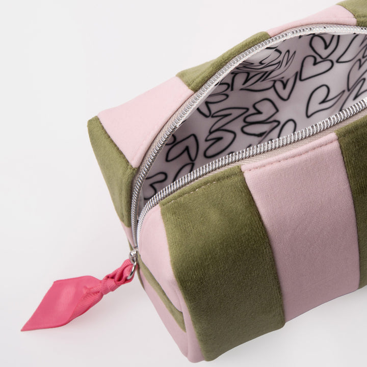 A close-up of a rectangular zippered pouch with alternating light pink and olive green velvet-like panels, a silver zipper, and a bright pink pull tab. The inside lining has a white background with black heart outlines.