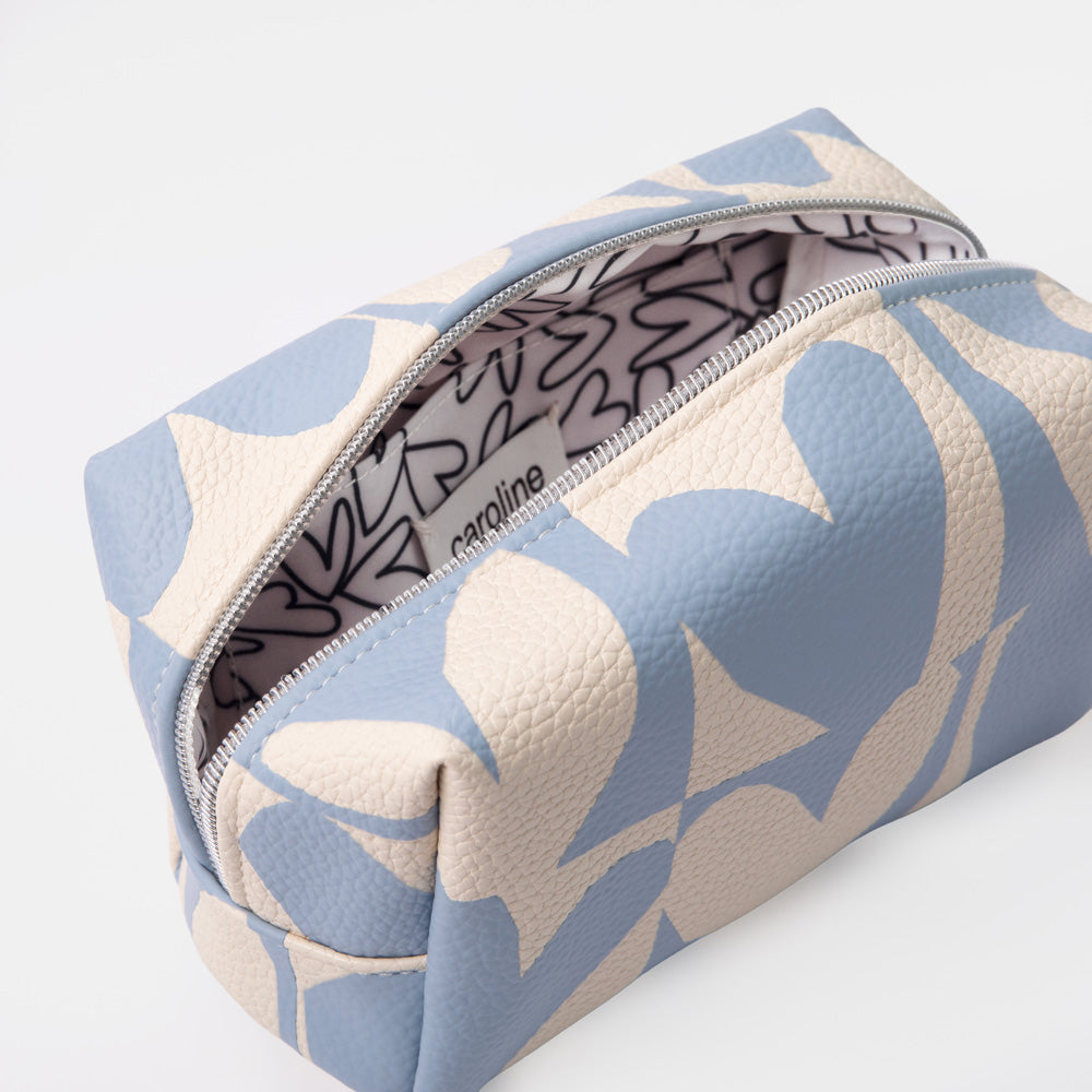 A rectangular zippered pouch with a textured pebbled finish, featuring a light blue and cream abstract floral pattern. The zipper is open, revealing a patterned interior with the brand name 'caroline' printed on it.