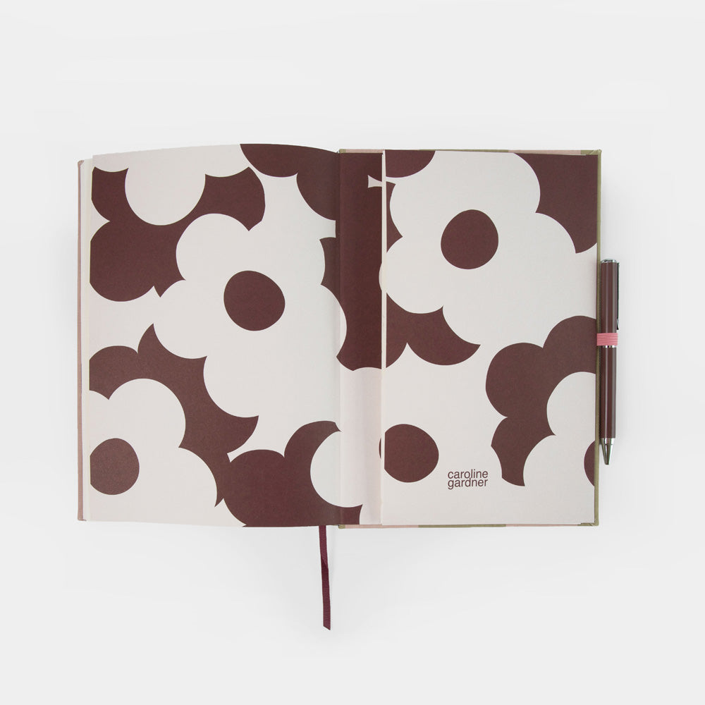 An open Caroline Gardner Journal with a brown and off-white floral pattern, featuring a matching pen in a pink loop on the right side.
