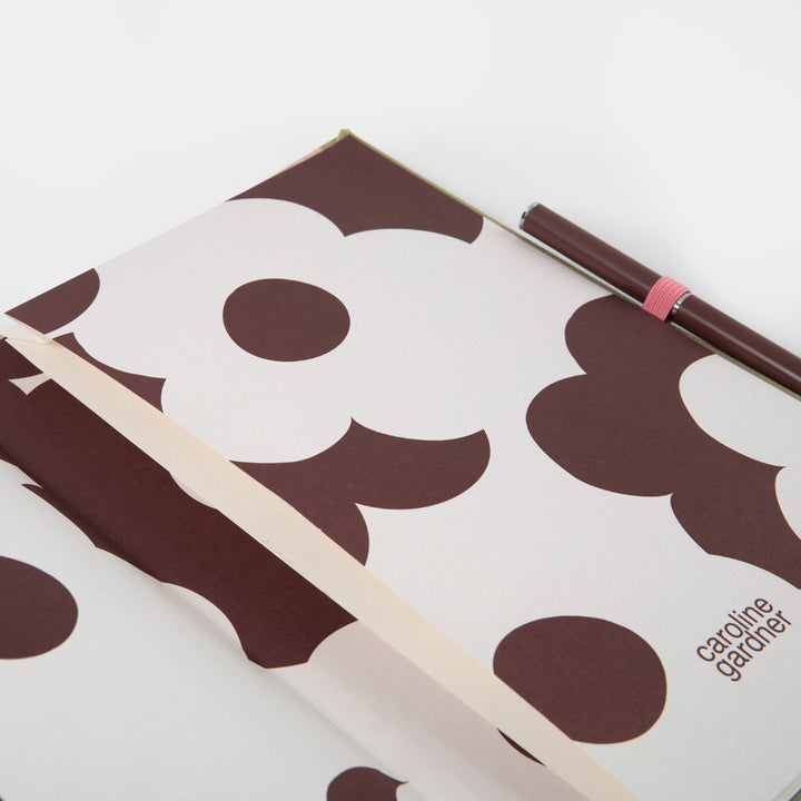 An open Caroline Gardner Journal with a brown and off-white floral pattern, featuring a matching pen in a pink loop on the right side.