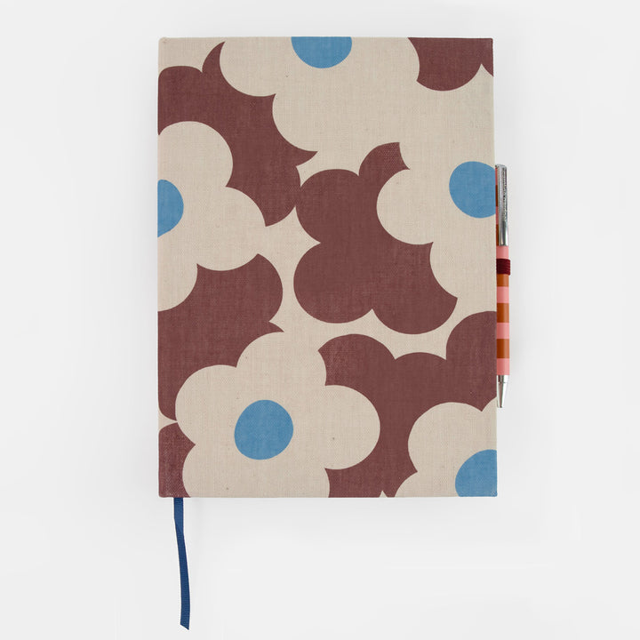 A fabric-covered notebook with a retro floral pattern in brown, cream, and blue, featuring a dark blue ribbon bookmark and a striped pen tucked into the side, set against a plain white background.