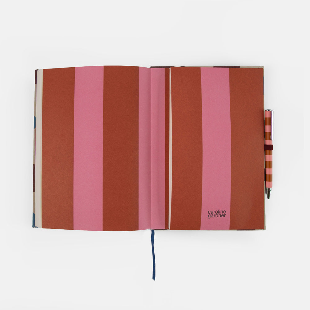An open notebook with vertical stripes in terracotta, dusty rose, and pink, shown on a clean white background with a matching striped pen tucked into the right side.