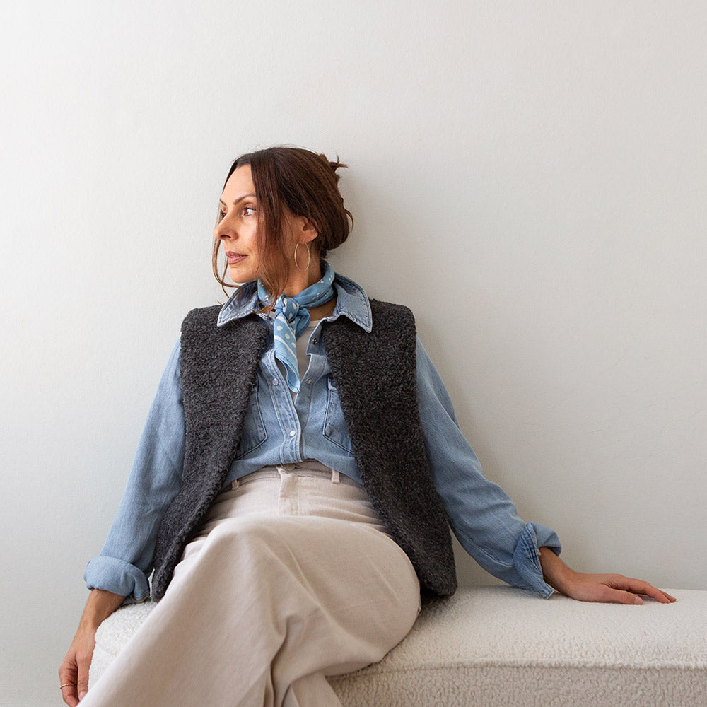 woman wearing a charcoal grey merino vest over a light wash denim shirt with cream trousers