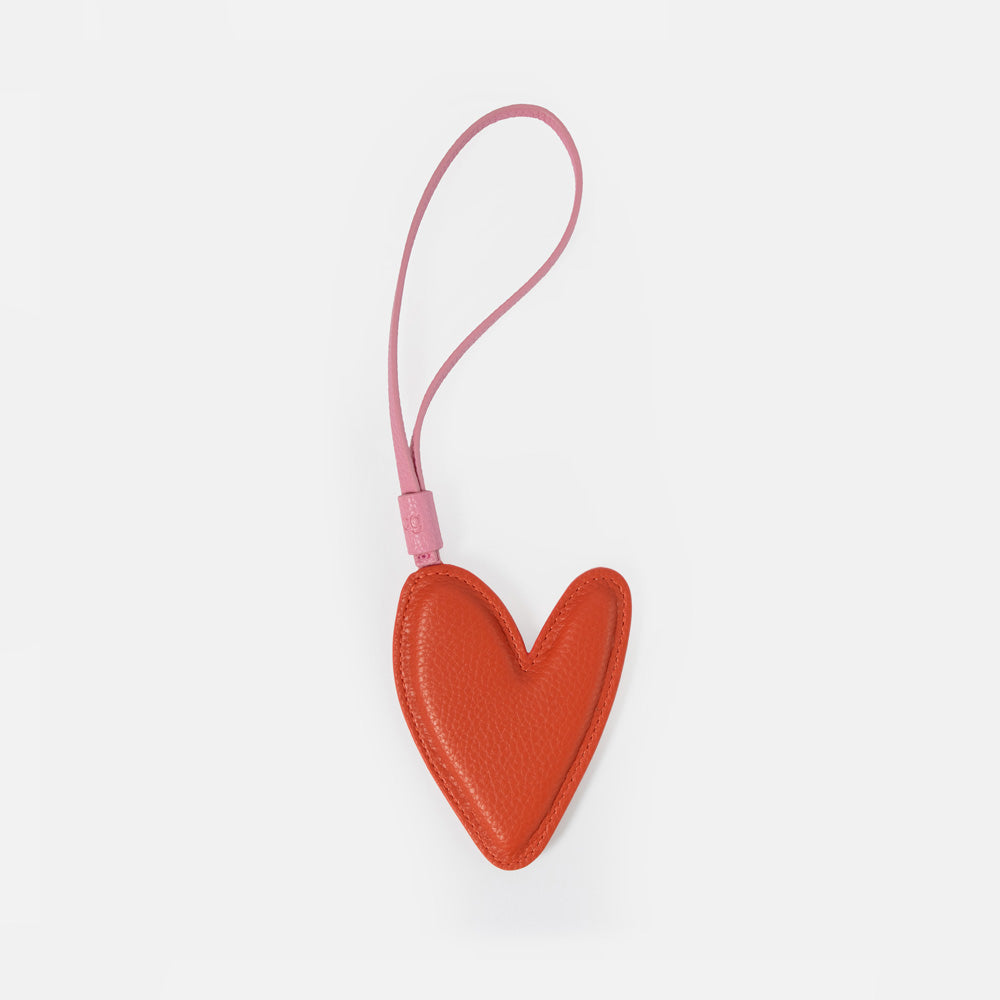 A small heart-shaped leather bag charm in a warm orange-red color with a matching pink loop strap, shown on a plain light gray background.