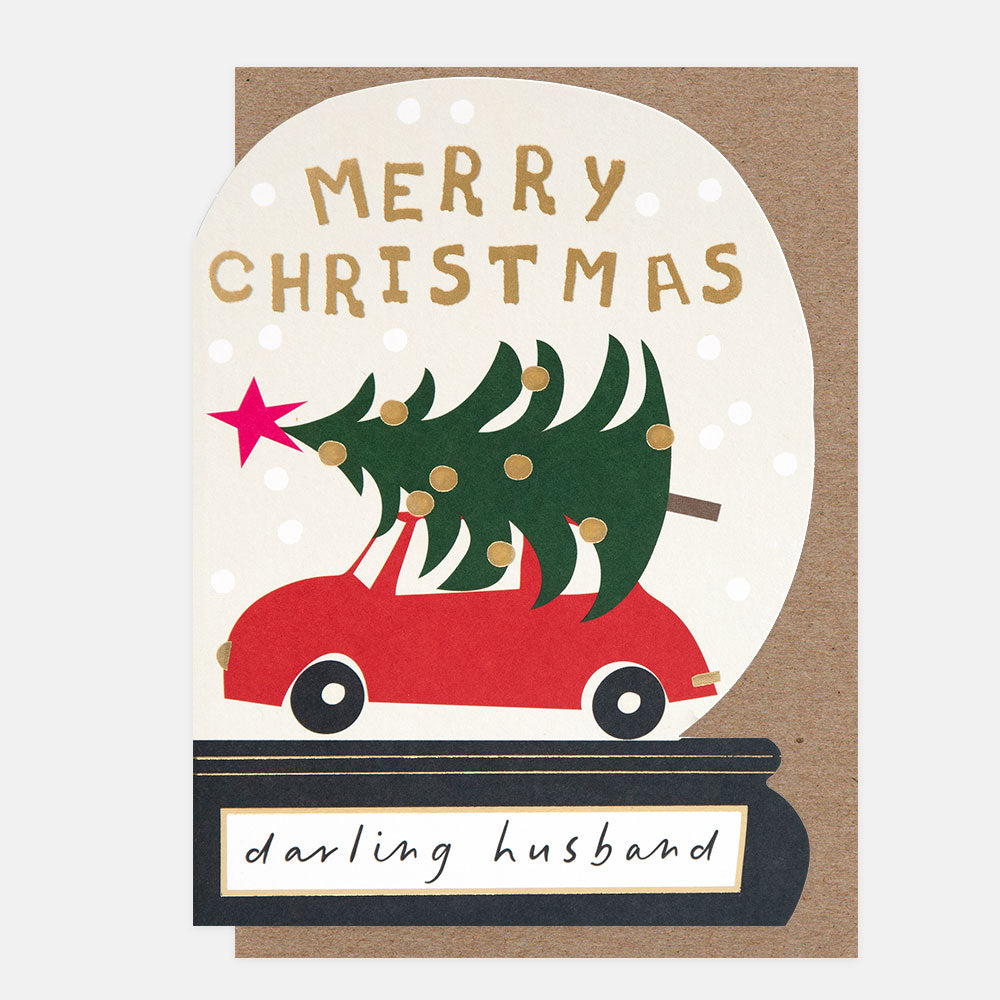 A Christmas greeting card with a kraft brown envelope behind it. The card shows a white oval snow globe illustration with the words “MERRY CHRISTMAS” in gold, a red car carrying a green Christmas tree, a pink star, and the phrase “darling husband” on a black base.