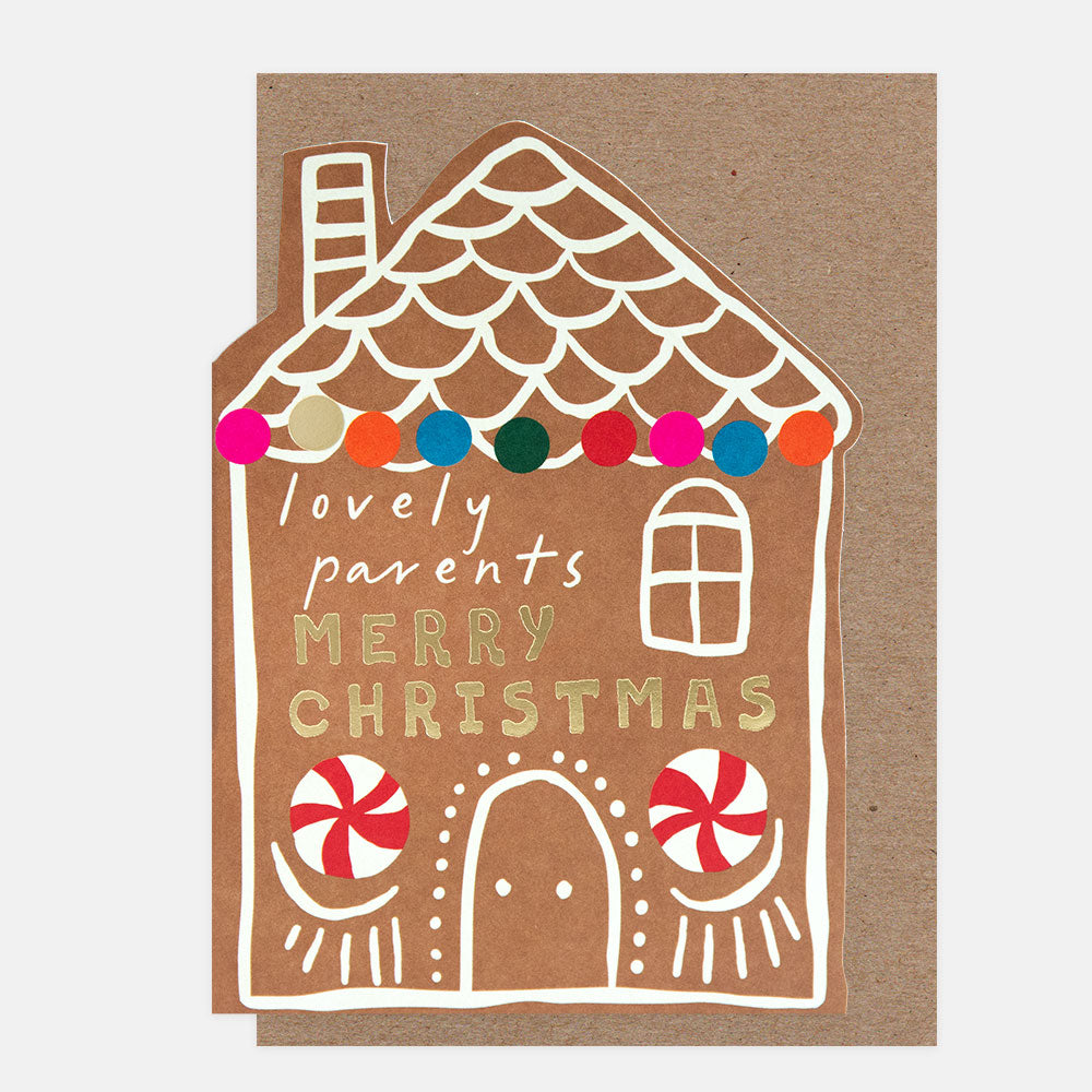 A gingerbread house shaped Christmas greeting card on a brown kraft envelope. The card features white icing-style outlines, gold foil text reading 'lovely parents MERRY CHRISTMAS', colorful string lights, and red-and-white peppermint candies on a brown background.