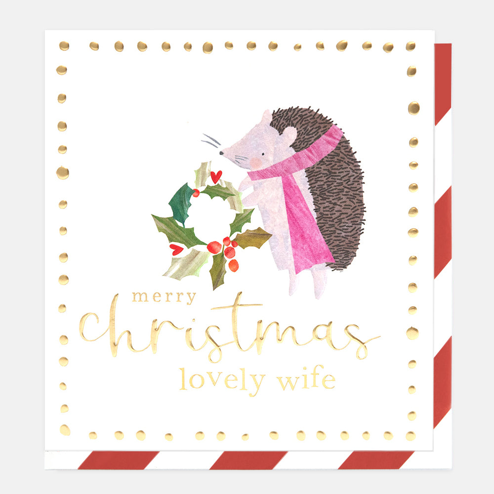 A square Christmas greeting card with a white background, gold dotted border, and a watercolor hedgehog wearing a pink scarf next to a wreath. The card reads 'merry christmas lovely wife' in gold script.