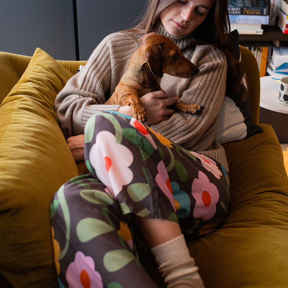 Woman wearing floral lounge pants and brown jumper holding a small brown dog on a yellow couch with a cosy atmosphere.