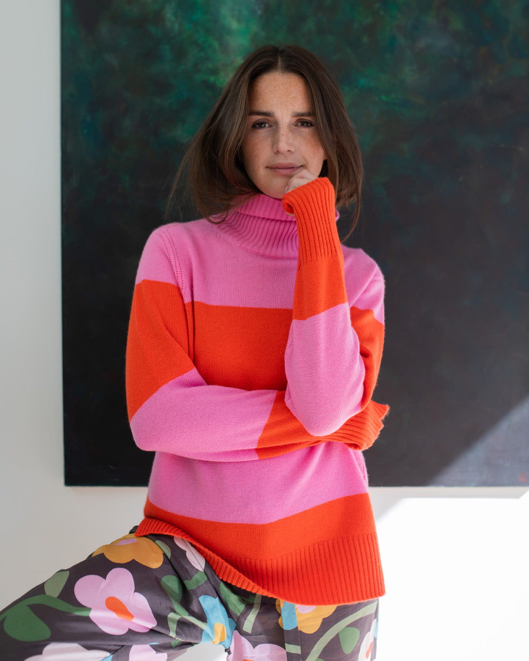 Woman wearing a pink and orange striped sweater with a colorful skirt against a dark abstract painting.