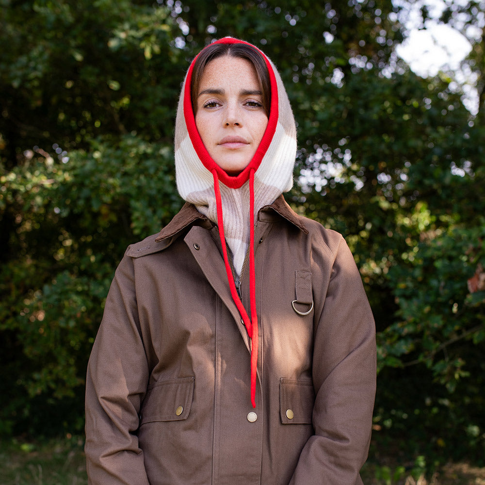 woman wearing a brown wax jacket and taupe & cream stripe knitted balaclava hood with a red trim and ties