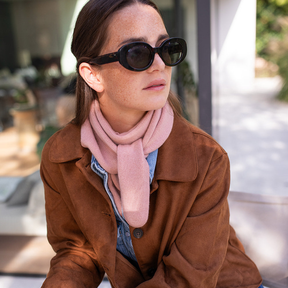 Woman wearing sunglasses and a brown jacket with a pink scarf outdoors