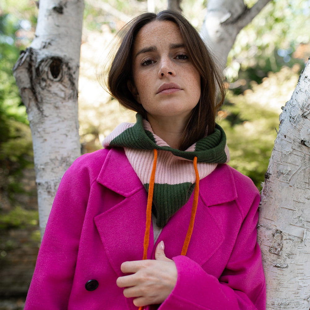 woman wearing a bright pink coat with a green and light pink striped balaclava hood with orange drawstrings