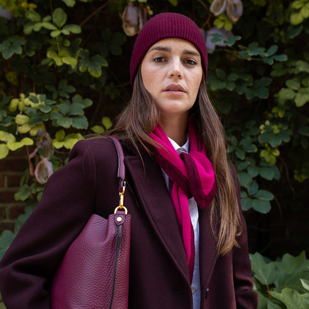 Woman wearing a purple coat, hat, and scarf standing against a green leafy background
