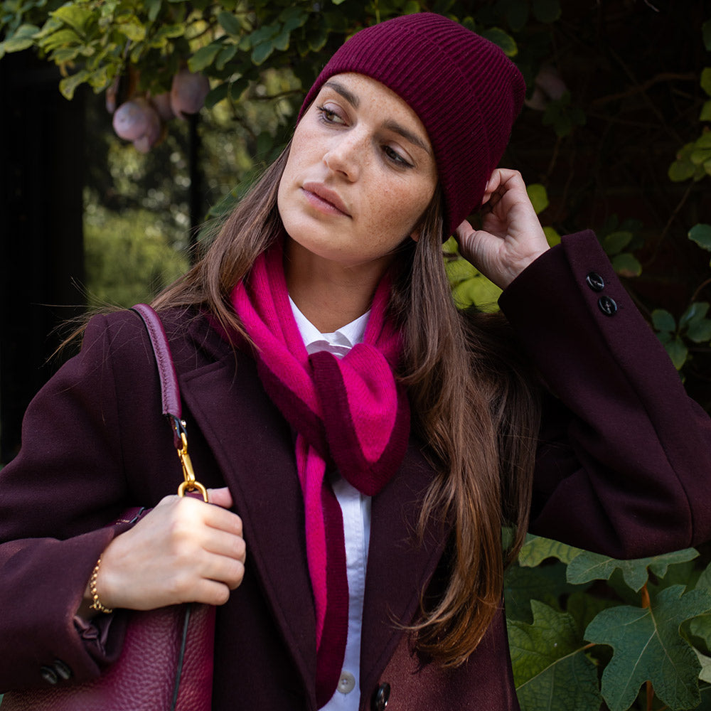 Woman wearing a burgundy coat, hat, and scarf outdoors with greenery in the background