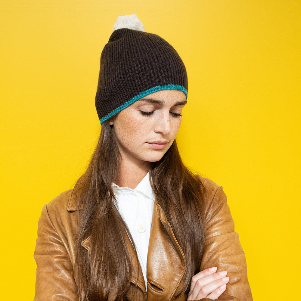 woman in front of a yellow backdrop wearing a tan leather jacket and dark grey beanie hat with a white bobble