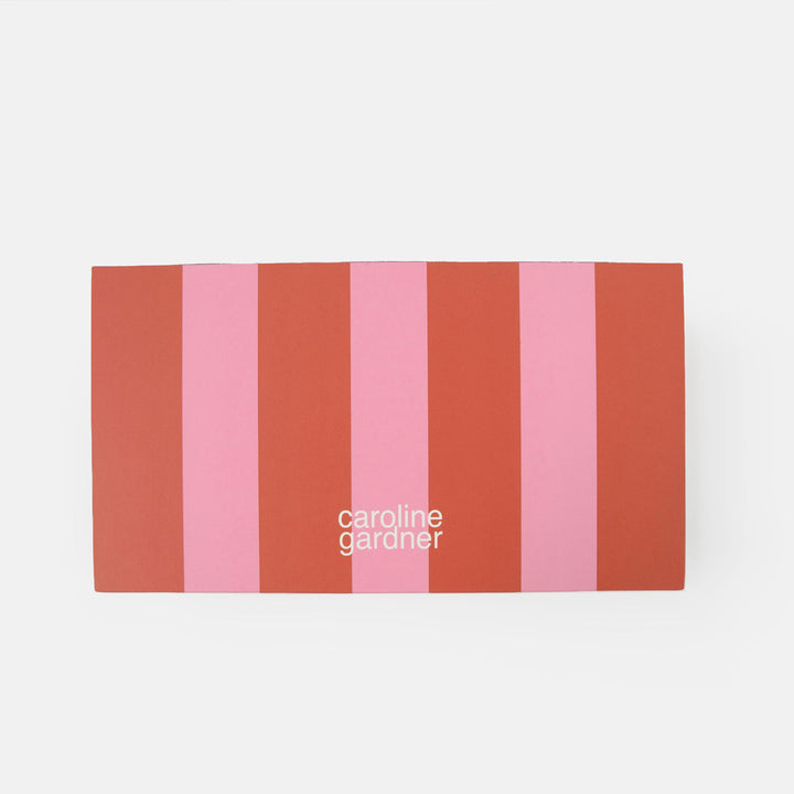 Desk pad reverse with vertical stripes in terracotta red, coral orange, and pink, featuring the white text 'caroline gardner' near the bottom center, set against a plain light gray background.