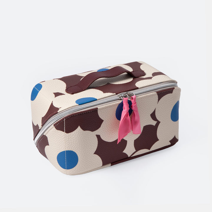 A rectangular zippered cosmetic bag with a cream background and large dark brown abstract shapes, accented with bright blue circles and a pink ribbon zipper pull, shown on a light gray background.