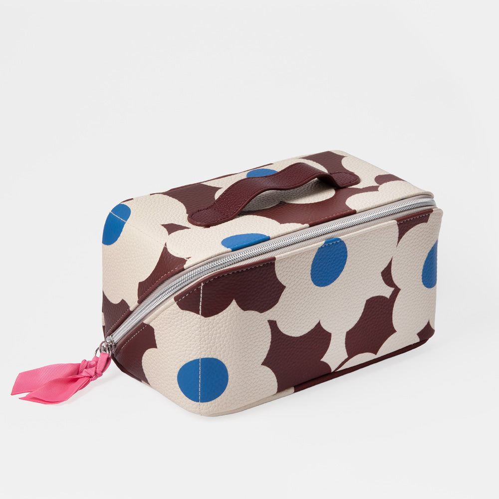 A rectangular zippered cosmetic bag with a cream background and large dark brown abstract shapes, accented with bright blue circles and a pink ribbon zipper pull, shown on a light gray background.