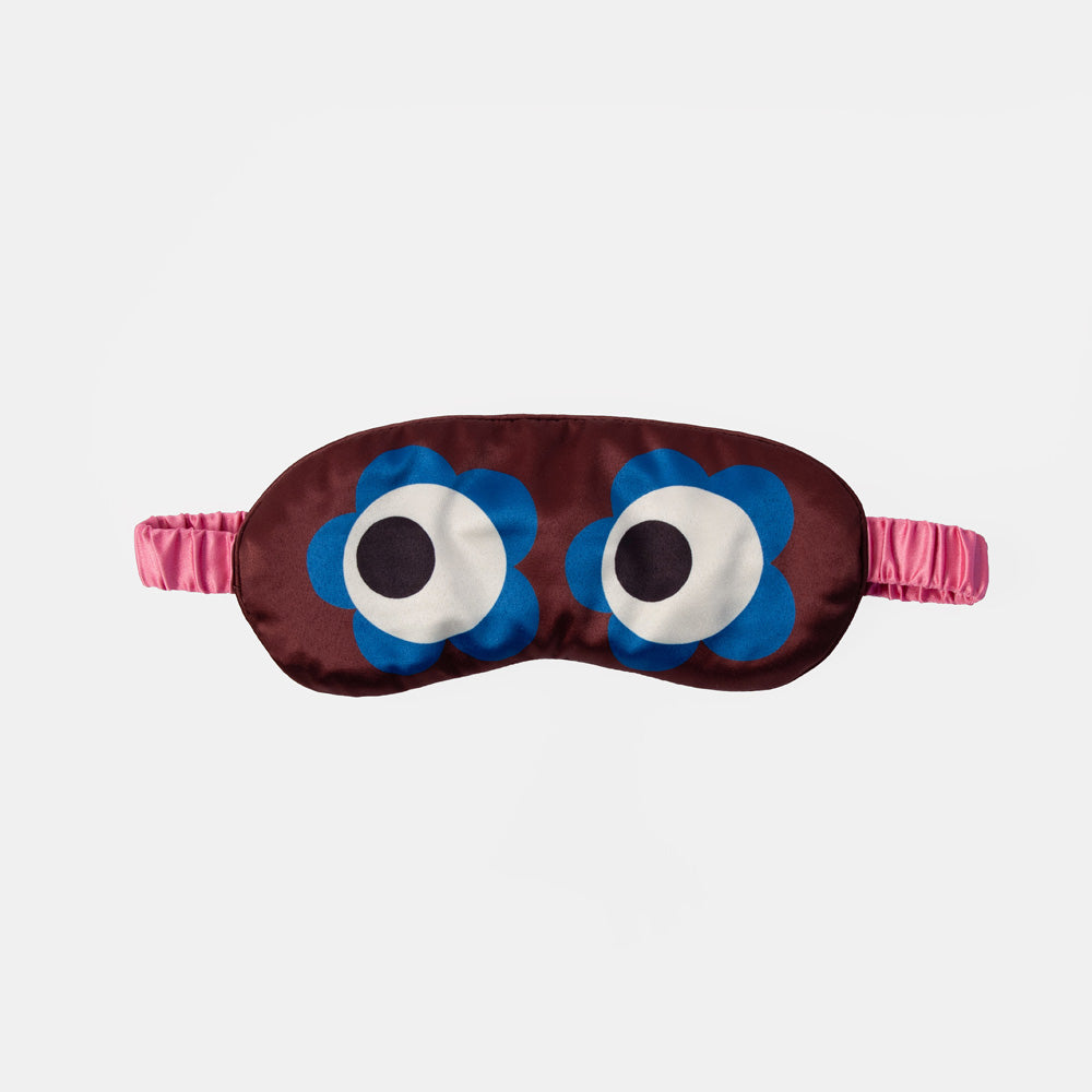 A novelty sleep mask with a dark brown satin body, featuring two large cartoon eyes with blue flower-shaped outlines, white sclera, and black pupils, with a pink elastic strap on a light gray background.