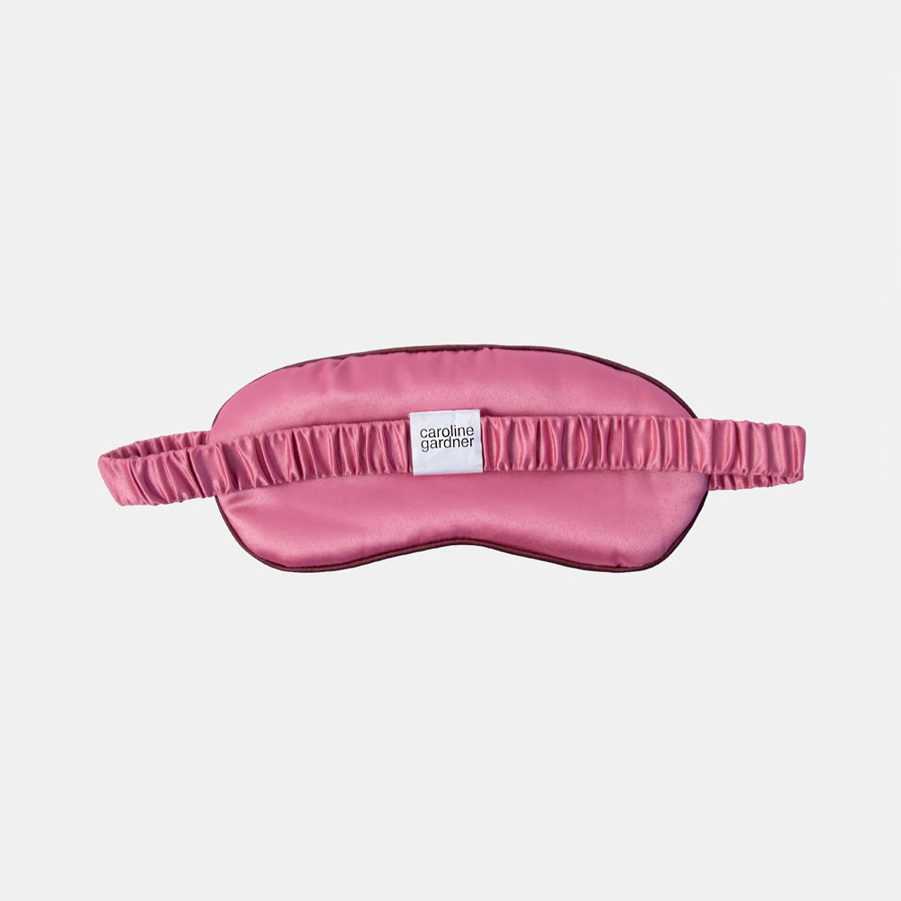 A pink satin sleep mask with an elastic strap, featuring a small white label that reads 'carole Gardner' in the center, set against a light gray background.